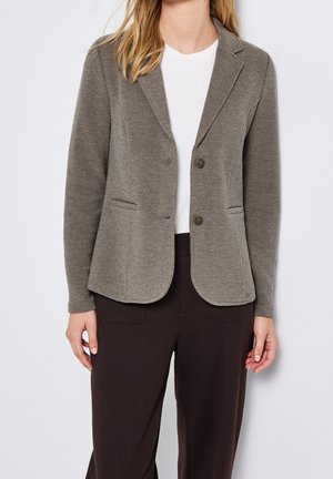 Femme portant un blazer gris texturé sur une chemise blanche et un pantalon foncé, debout devant un fond clair uni.