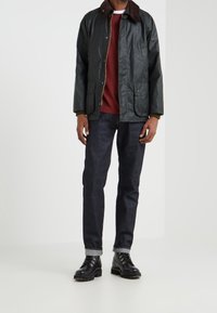 Tmavě zelená nepromokavá bunda s hnědým manšestrovým límcem, dvojitými předními kapsami, nošená přes červený svetr a tmavé d�žíny, doplněná černými botami.