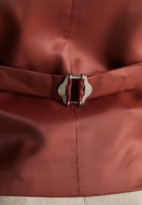 Brauner Satinstoff mit glatter Textur, der in der Mitte eine metallische rechteckige Schnalle aufweist, gesichert durch einen passenden Gürtel aus dem selben Material.