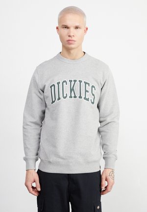 Sudadera gris con el texto verde "DICKIES", cuello y puños acanalados, textura suave y corte holgado, llevada sobre pantalones cargo negros.