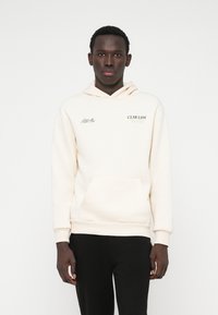 Κρέμα φούτερ από βαμβάκι με μπροστινή τσέπη, κουκούλα με κορδόνια και εκτυπωμένα λογότυπα σε μαύρο και πορτοκαλί. Φορεμένο με μαύρο παντελόνι.