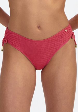 Czerwone, teksturowane majtki od bikini z wiązaniami po bokach i złotymi metalowymi dodatkami, noszone przez kobietę, widoczna środkowa część ciała.