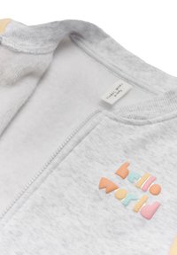 Prenda gris con cremallera que presenta una textura suave, con el texto "hola mundo" en colores pastel en la parte frontal.