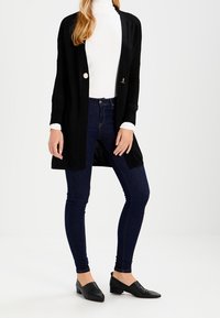 Schwarzer lange Cardigan mit einem einzelnen Knopf, getragen über einem weißen Rollkragenpullover; kombiniert mit dunkelblauen engen Jeans und schwarzen Ballerinas.