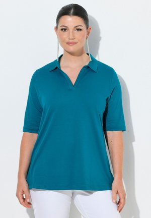 Kobieta o ciemnych włosach z niskim kokiem, nosząca długie srebrne kolczyki, turkusową koszulę z krótkim rękawem i kołnierzykiem oraz białe spodnie, stoi na tle białej ściany.