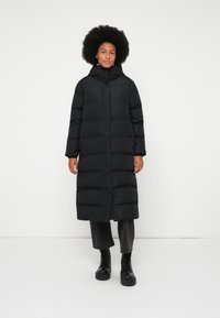 Kvinne med afrofrisyre iført en lang svart polstret jakke, svarte cropped bukser og svarte snørestøvler som står mot en enkel hvit bakgrunn.