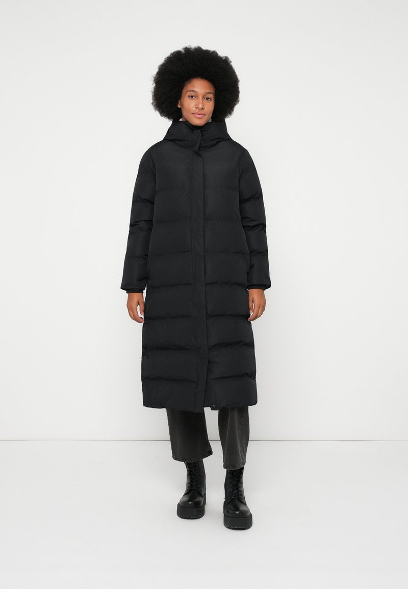 Kvinne med afrofrisyre iført en lang svart polstret jakke, svarte cropped bukser og svarte snørestøvler som står mot en enkel hvit bakgrunn.