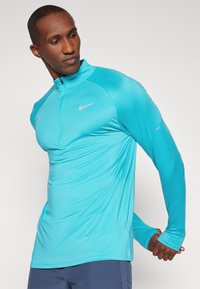 Uomo che allunga un braccio dietro la schiena indossando una maglietta atletica a maniche lunghe blu brillante e pantaloni corti blu navy su uno sfondo uniforme.