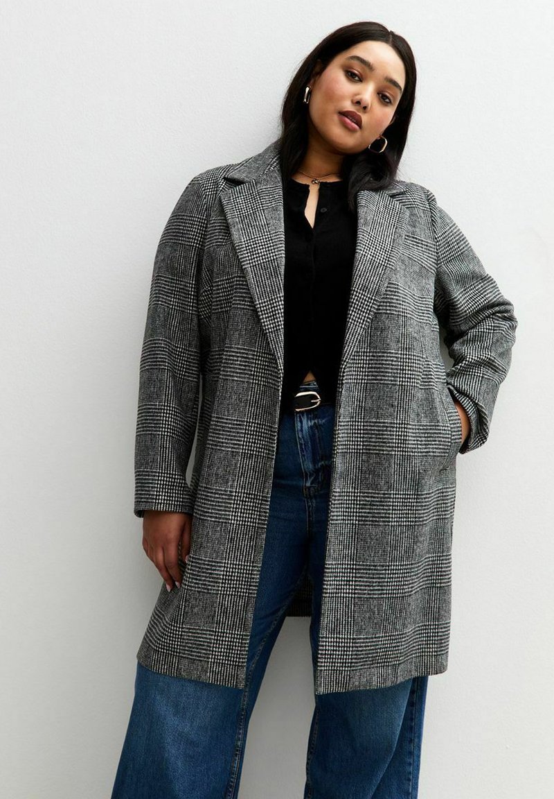 Cappotto oversize a quadri grigi con colletto, tasche frontali e maniche lunghe, indossato sopra una maglia nera e jeans larghi blu.
