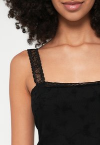 Mulher a usar um top sem mangas preto em renda com padrões florais, mostrando o decote, o ombro e parte do cabelo encaracolado.