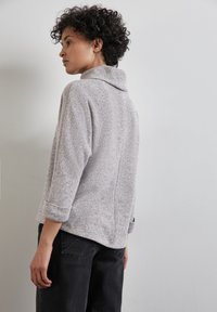 Grauer, strukturierter Pullover mit hohem Kragen und hochgerollten Ärmeln, kombiniert mit schwarzen, schmal geschnittenen Hosen. Hat einen lockeren Schnitt und einen strukturierten Rücken.