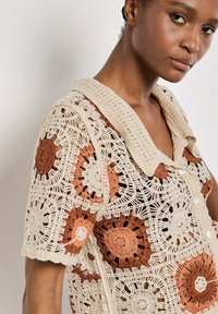 Häkelhemd mit kragenförmigem Design in Beige mit orangefarbenen und braunen Blumenmustern. Verfügt über Knöpfe und eine filigrane, spitzenähnliche Textur.