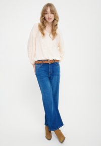 Lichtroze blouse met kanten details, gecombineerd met blauwe uitlopende jeans en tan enkellaarsjes. Model staat tegen een witte achtergrond.