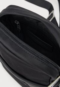 Μαύρη τσάντα ώμου από ύφασμα με κλείσιμο με φερμουάρ, εσωτερική τσέπη και υφή επένδυσης. Περιλαμβάνει ένα επίπεδο λογότυπο στο πλάι.