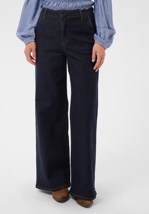 Personne portant un jean bleu foncé à jambes larges, un chemisier bleu pâle à manches longues et des chaussures marron, debout devant un fond uni.