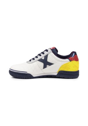 Zapatilla blanca con cordones negros, suela negra, logotipo "X" azul marino, parche amarillo en el talón y detalle rojo en la parte trasera.