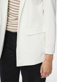 Valkoinen bleiseri, jossa on kaksi etupussia, yhdistetty beige-valkoraidalliseen toppiin. Mustat tekstuuriset housut näkyvät alaosasta.