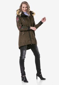 Olivgrüne Parka mit einer Kunstpelzkapuze, floralem Stickmuster auf den Ärmeln, mit Reißverschlusstaschen und schwarzen Akzenten; kombiniert mit engen schwarzen Jeans und hohen Schuhen.