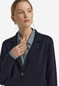 Marineblaue Strickjacke mit strukturierter Masche, Knopfverschluss und Reverskragen, kombiniert mit einer hellblauen, gestreiften Bluse darunter.