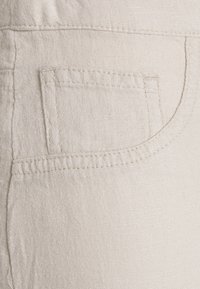 Beżowy detal kieszeni z tkaniny z widocznym przeszyciem na teksturowanym materiale lnianym, z zaokrąglonymi krawędziami i delikatnymi wzorami słojów.