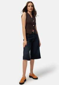Top marrón sin mangas con escote en V y dos botones dorados, combinado con culottes de mezclilla azul oscuro y mocasines de ante naranja con suelas gruesas.