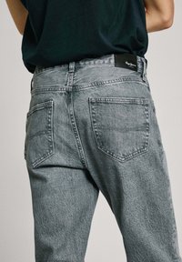 Jeans de mezclilla gris con un diseño clásico de cinco bolsillos y una textura ligeramente desgastada. Etiqueta de cuero negra en la cinturilla, corte recto.