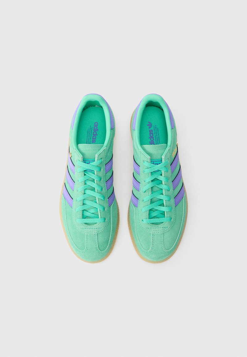 Green suede sneakers feature purple stripes and laces, with a textured upper and a gum rubber sole, showcasing a minimalist design.
