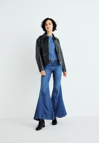 Zwarte leren cropped jack over een blauwe denim top en uitlopende blauwe jeans, gedragen met zwarte enkellaarsjes. Soepele textuur en een getailleerde pasvorm.