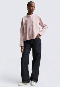 Ljusrosa randig skjorta med knappar och en bröstficka, kombinerad med mörkblå vida jeans och vita sneakers.