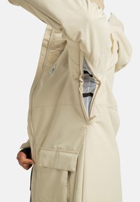 Beżowa kurtka z zamkiem błyskawicznym w okolicy pach, z czarno-białą siateczkową podszewką, kieszenią z klapką i ręką osoby blisko dolnej części.