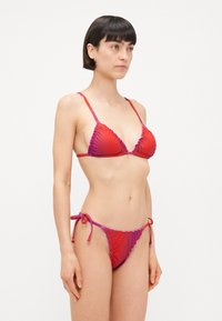 Bikini set u crveno-ružičastom gradijentu s volanastim rubovima, tankim podesivim naramenicama i donjim dijelom na vezanje. Glatka, rastezljiva tkanina.