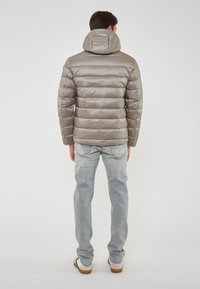 Uomo che indossa una giacca trapuntata marrone chiaro con cappuccio, jeans azzurri e sneaker bianche, in piedi di spalle su uno sfondo bianco.