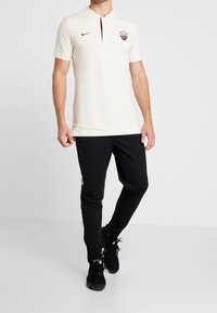 Jasna kremowa koszulka polo z krótkimi rękawami, czerwonymi akcentami i kołnierzykiem. Połączona z czarnymi zwężanymi spodniami sportowymi i czarnymi trampkami.