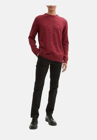 Pull en maille texturée rouge avec un col rond, associé à un pantalon noir et des chaussures noires. Le tissu présente un motif côtelé subtil.