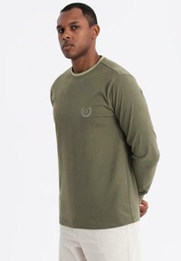 Olivenfarbene langärmelige T-Shirt mit Rundhalsausschnitt und subtiler Logodetail auf der Brust. Hergestellt aus weichem, leichtem Baumwollmaterial.