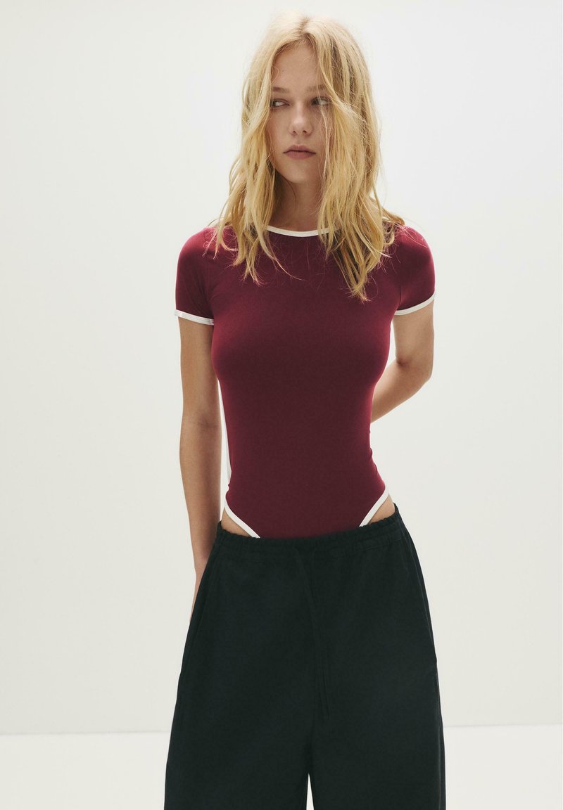 Donna bionda con capelli sciolti, vestita con un body aderente color bordeaux con elementi bianchi e pantaloni neri larghi, in piedi contro uno sfondo chiaro e semplice.