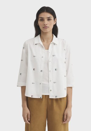 Woman with long dark hair wearing a white button-up shirt with small book prints over a white top and brown pants, standing against plain background.