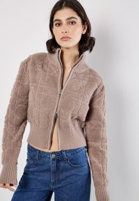 Suéter con cremallera recortado en color topo, que presenta una tela texturizada con un patrón geométrico, puños acanalados y un cuello alto.
