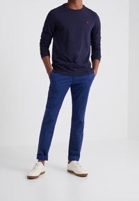 Camicia a maniche lunghe blu navy con un piccolo logo rosso, abbinata a pantaloni blu e sneakers bianche con dettagli marroni. Design semplice e vestibilità casual.