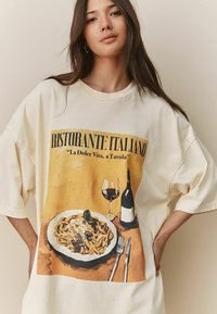 Crème oversized T-shirt featuring a graphic print van een Italiaans restauranttafereel met pasta, wijnglas en gedetailleerde tekst in bold zwart.