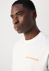 Balta crewneck džemperis su raukšlėta apykakle, su oranžiniu logotipu, kuriame užrašyta "by MarcoPoloDenim" krūtinėje. Lygus paviršius.
