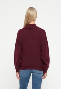 Burgundská oversized mikina s vysokým ribovaným golierom a balónovými rukávmi, textúrovaná tkanina s jemným povrchom, skombinovaná s modrými džínsami.