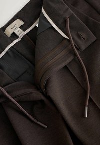 Pantaloni marroni con vita regolabile, tasche con zip e una consistenza morbida. Etichetta indica il marchio NEXT e la taglia 34R.