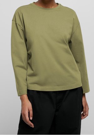Person trägt ein olivgrünes Langarmshirt und schwarze Hose, steht vor einem neutralen grauen Hintergrund.
