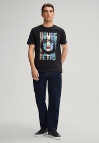 Zwarte t-shirt met een retro graphic print met kleurrijke gezichten en de tekst "ROUGE RETRO," gecombineerd met donkere spijkerbroeken en zwarte sneakers.