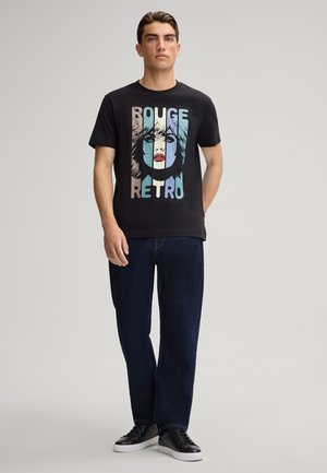 Schwarzes T-Shirt mit einem Retro-Grafikdruck, der bunte Gesichtillustrationen und den Text "ROUGE RETRO" zeigt, kombiniert mit dunklen Jeans und schwarzen Sneakers.