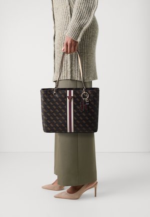 Person der holder en brun monogrammeret Guess håndtaske med stribet accent, iført en strikket cardigan, olivenfarvet nederdel og beige hæle.