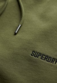 Sudadera verde oliva de tejido suave, con capucha, que presenta un logo bordado en negro de "SUPERDRY" en el frente. Acabado texturizado.