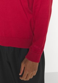 Roter Strickpullover mit gerippten Bündchen und "HUG"-Logo. Nahaufnahme des Ärmels und der Hand, die Textur und Passform über schwarzen Hosen zeigt.