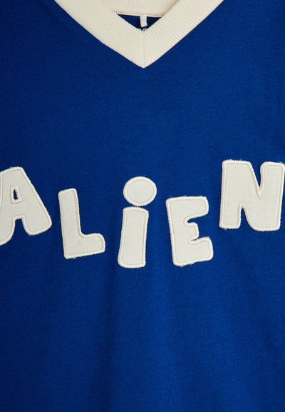 Μπλε μπλούζα με λαιμόκοψη σε σχήμα V που δείχνει λευκά ραμμένα γράμματα με την επιγραφή "ALIEN" στο στήθος.